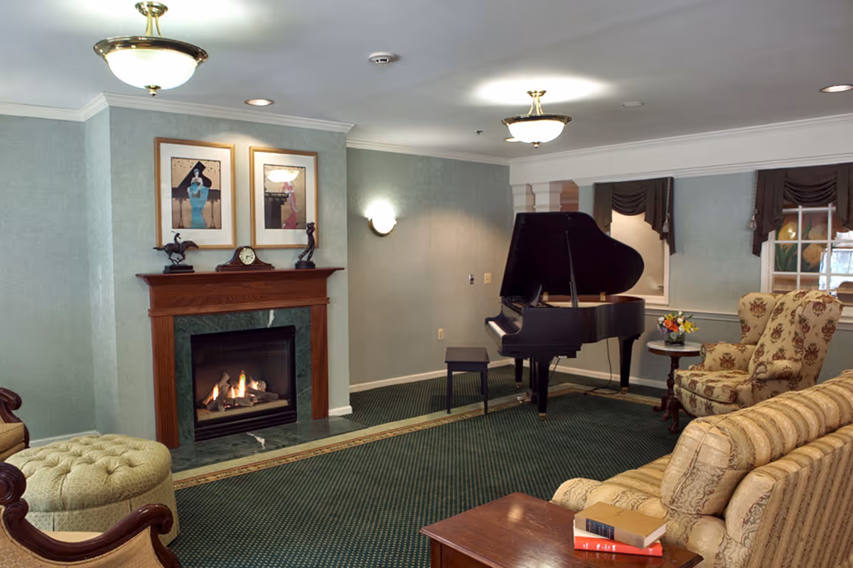 A cozy living room area featuring a lit fireplace with a wooden mantle, two framed artworks above it, a grand piano with a bench, a floral upholstered armchair, a striped sofa, a round side table with a flower arrangement, and a wooden coffee table with books on it. The room has green carpet, light green walls, and ceiling lights.