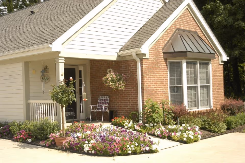 Exterior view of a single-story senior living facility unit with a brick and white siding facade. The entrance features a small porch with a chair, hanging flower basket, and potted plants. The front yard is landscaped with colorful flowers and shrubs.