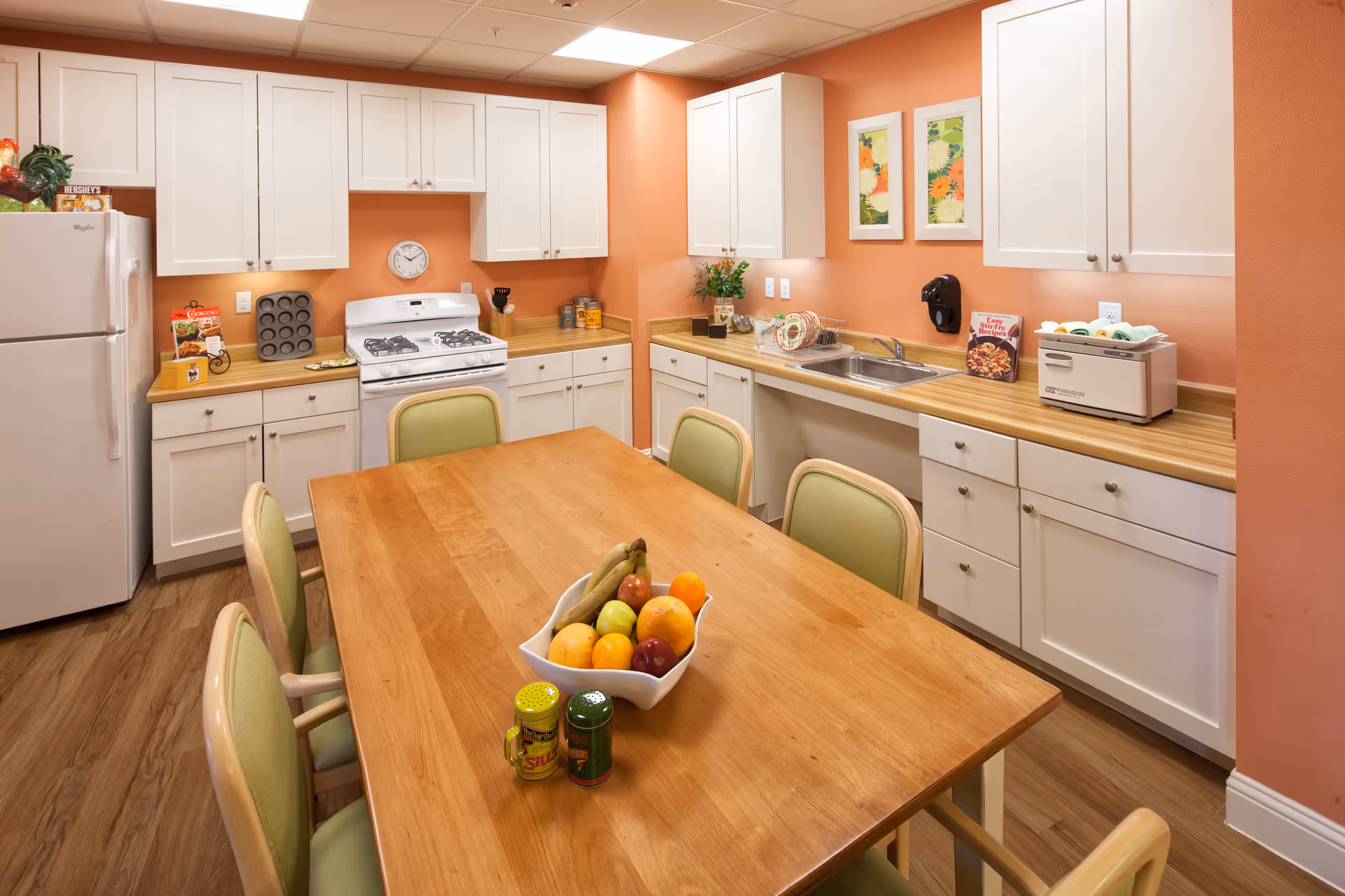 A bright communal kitchen with a wooden table and fruit bowl, green chairs, white cabinets, a stove, sink and refrigerator against coral-colored walls.
