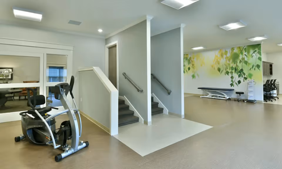Interior view of a senior living facility showing a recumbent exercise bike on the left, a staircase with handrails in the center, and a treatment or therapy area with a medical examination table and stools on the right. The wall behind the therapy area features a mural of green and yellow leaves.