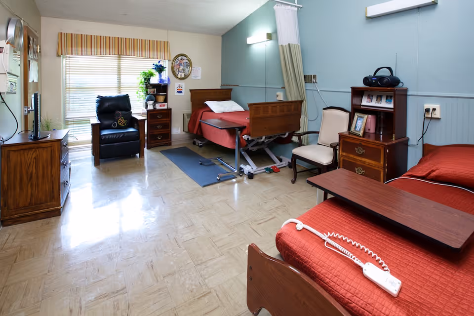 Shared senior care bedroom with two red-covered beds, an adjustable hospital bed, recliner, wooden furniture and a television.