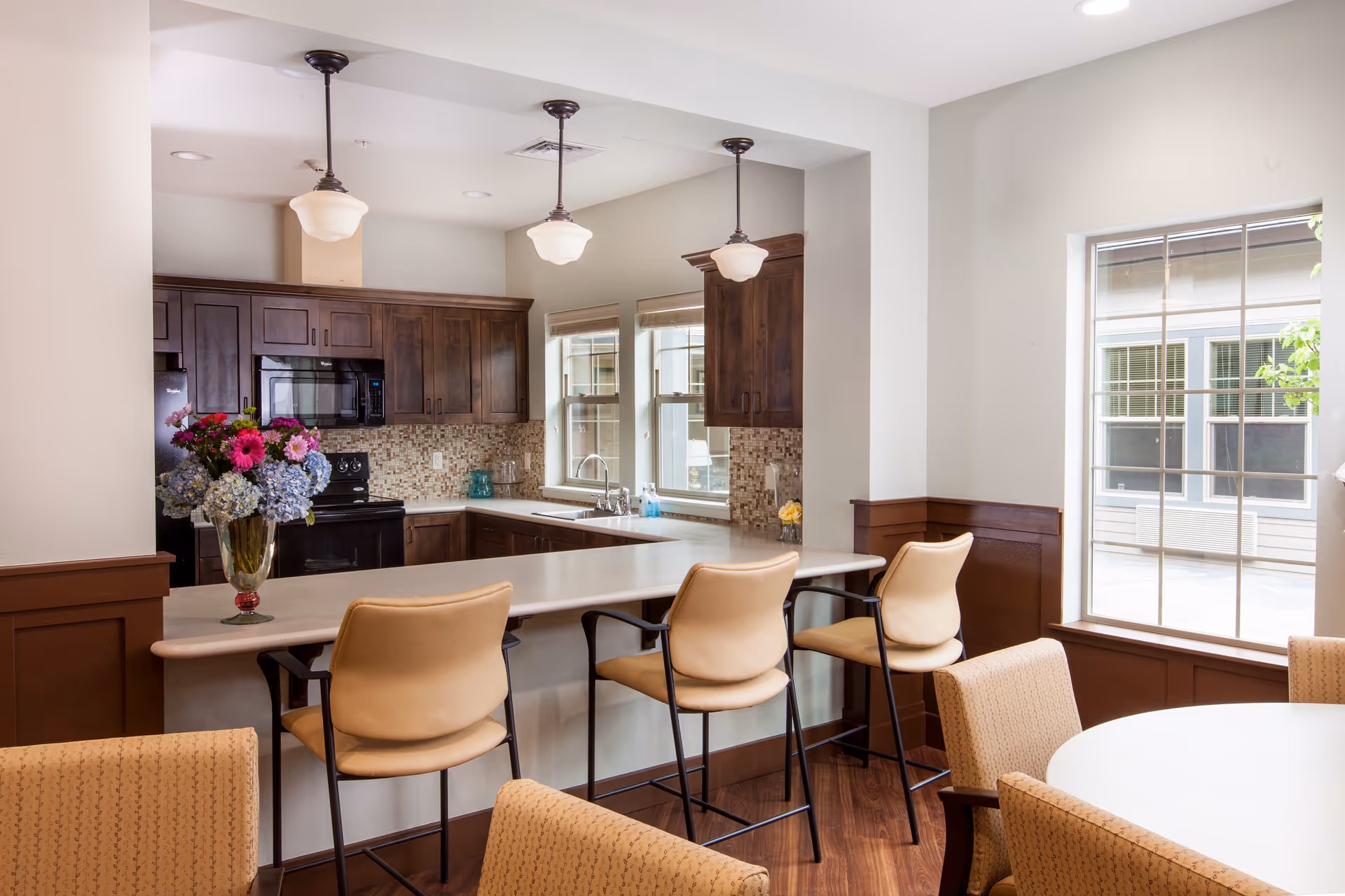 A bright kitchen area with dark wooden cabinets, a tiled backsplash, and a countertop with three beige bar stools. A vase with colorful flowers is placed on the counter. Adjacent to the kitchen is a dining area with a round table and upholstered chairs, next to a large window letting in natural light.