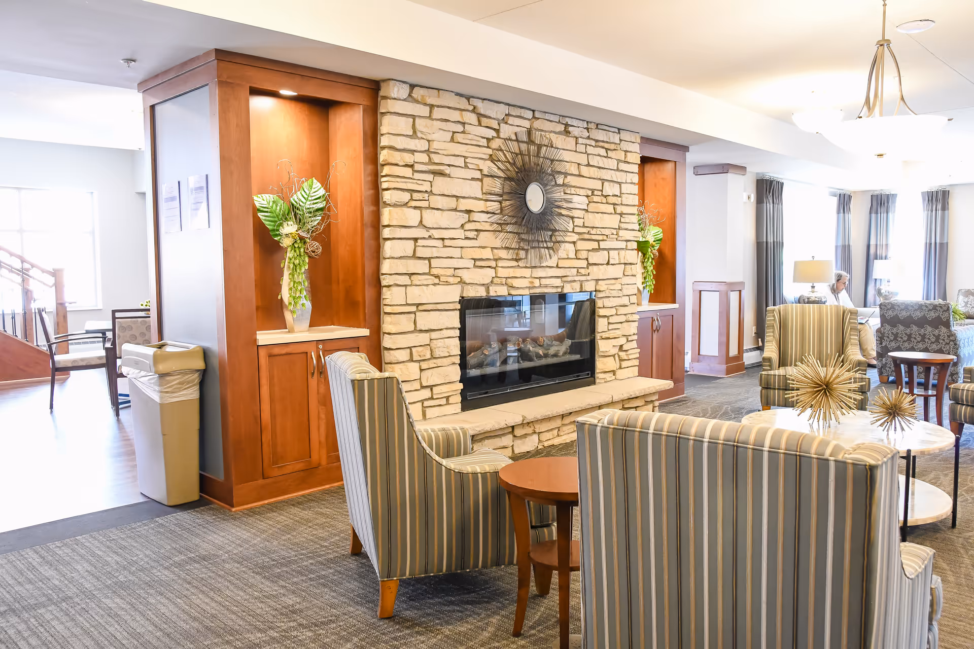 A bright and cozy living room area in a senior living facility featuring a stone fireplace with a decorative sunburst mirror above it. The room has striped armchairs arranged around a small round table with decorative gold starburst sculptures. There are built-in wooden shelves with vases of green plants on either side of the fireplace. In the background, a person is seated near windows with curtains, and the space is well-lit with ceiling lights and lamps.