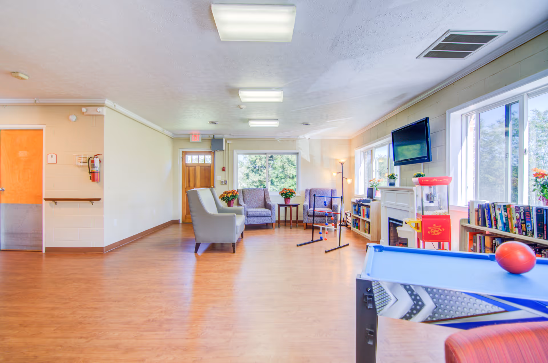Bright common lounge in a senior living facility with seating, bookshelves, a wall-mounted TV, and a game table by large windows.