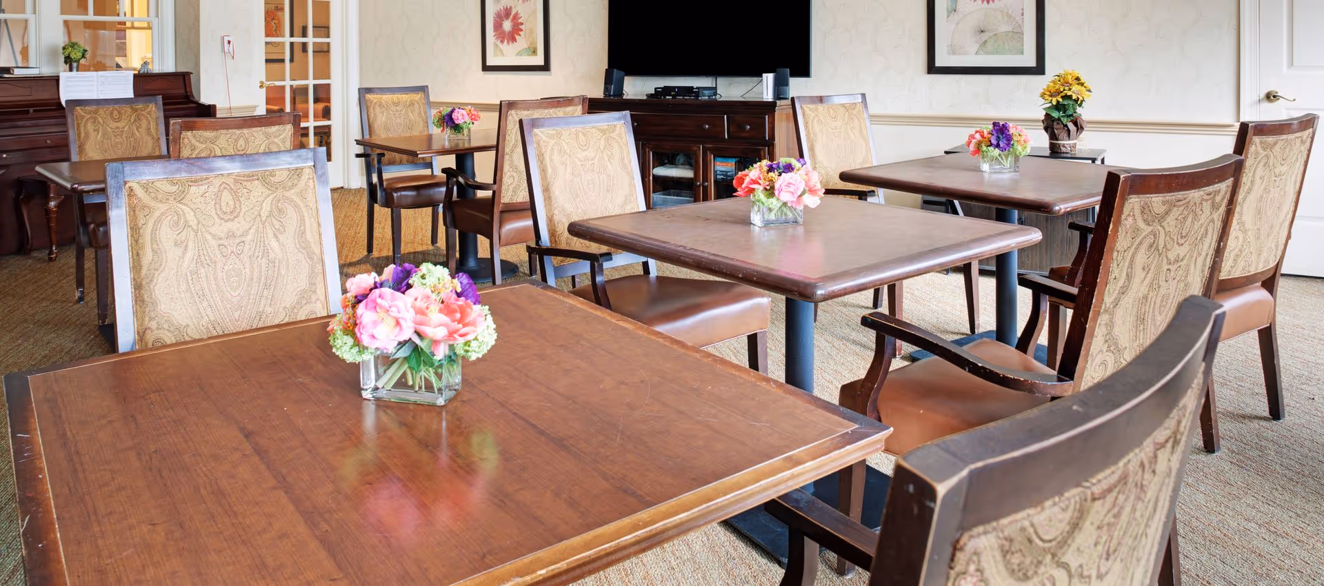 A bright dining room with wooden tables and upholstered chairs, small floral centerpieces, a TV and piano in the background.