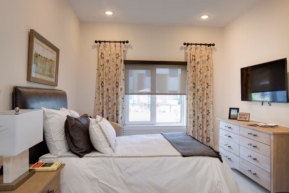 A neatly made bed with white and dark pillows in a senior living bedroom. The room features a window with floral curtains and a roller shade, a wooden dresser with framed photos and an open book, a wall-mounted flat-screen TV, a bedside table with a lamp and a book titled 'Pride and Prejudice', and a framed landscape painting on the wall.