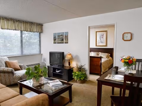 Comfortable living room with a sofa, armchair, coffee table, TV on a stand, dining table, plants, and a doorway showing a bedroom.
