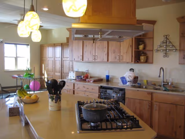 A bright kitchen with wooden cabinets and a central island featuring a gas stove with a pot on it. The kitchen has hanging pendant lights, a sink with dishes drying in a rack, and various kitchen utensils and appliances on the countertops. A window lets in natural light, and there is a fruit bowl with bananas and other fruits on the island.