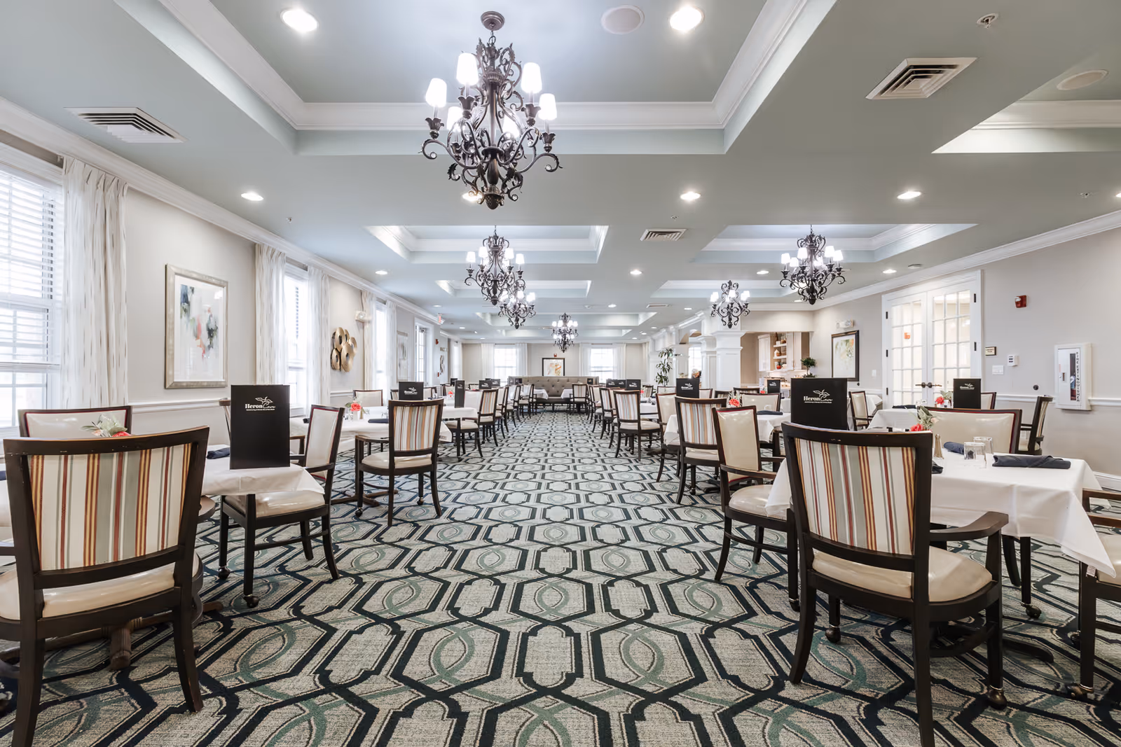 A spacious dining room with multiple tables and chairs arranged neatly. The room features patterned carpet flooring, elegant chandeliers hanging from the ceiling, large windows with white curtains allowing natural light, and light-colored walls adorned with framed artwork. Each table has menus labeled 'Heron Cove Assisted Living & Memory Care'.