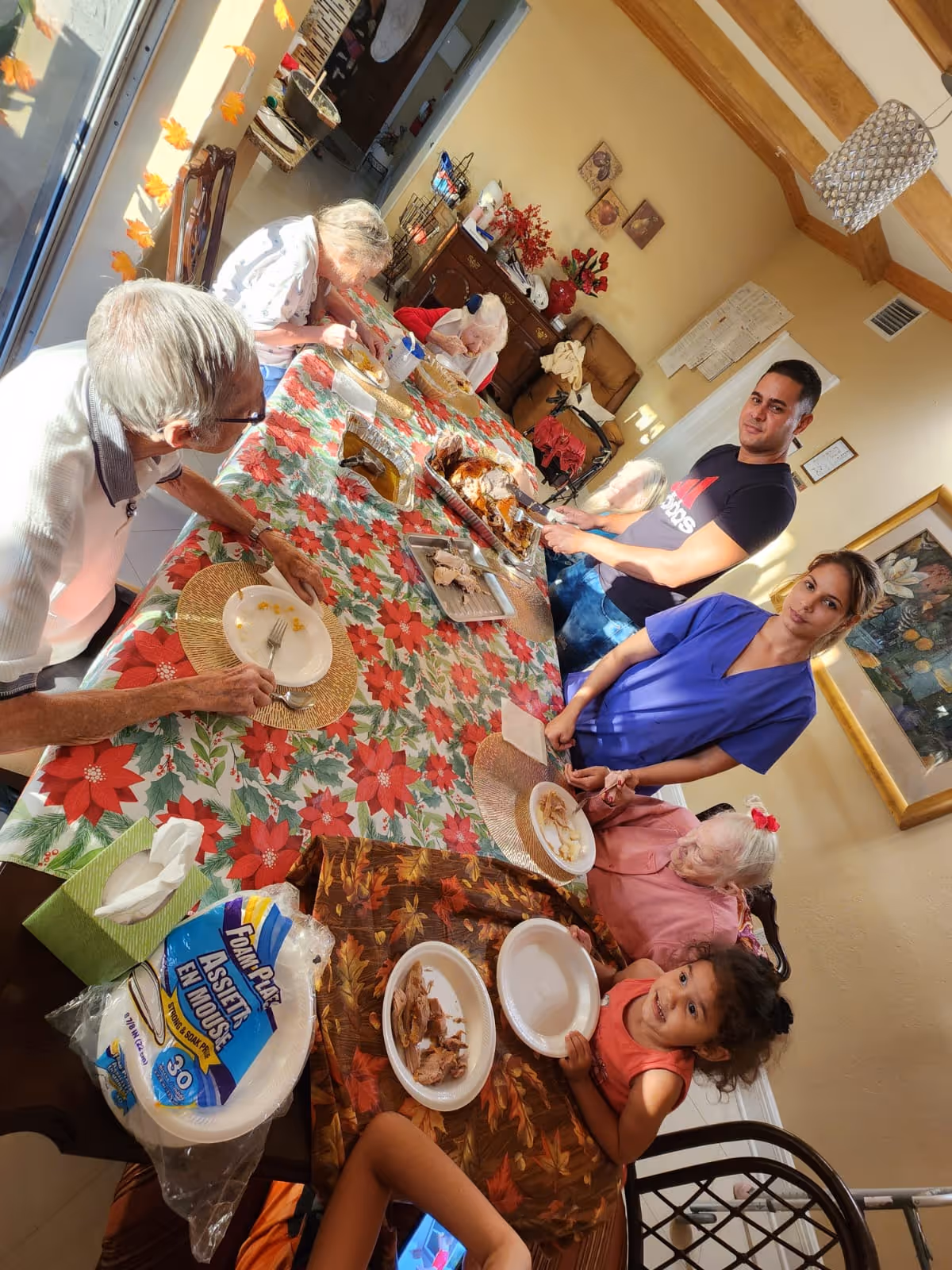 A group of elderly people and two caregivers gathered around a dining table covered with a festive red and green poinsettia tablecloth. The table has plates with food remnants, a tray of carved meat, and disposable plates and napkins. The room is warmly lit with beige walls, wooden ceiling beams, and a chandelier. A young child is also seated at the table, looking at the camera.