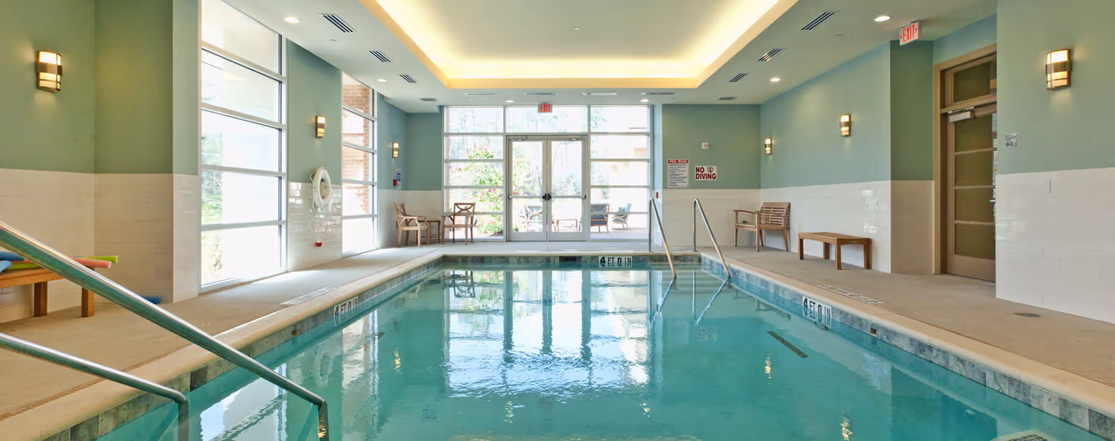 Bright indoor swimming pool area with large windows, seating benches, and metal handrails.