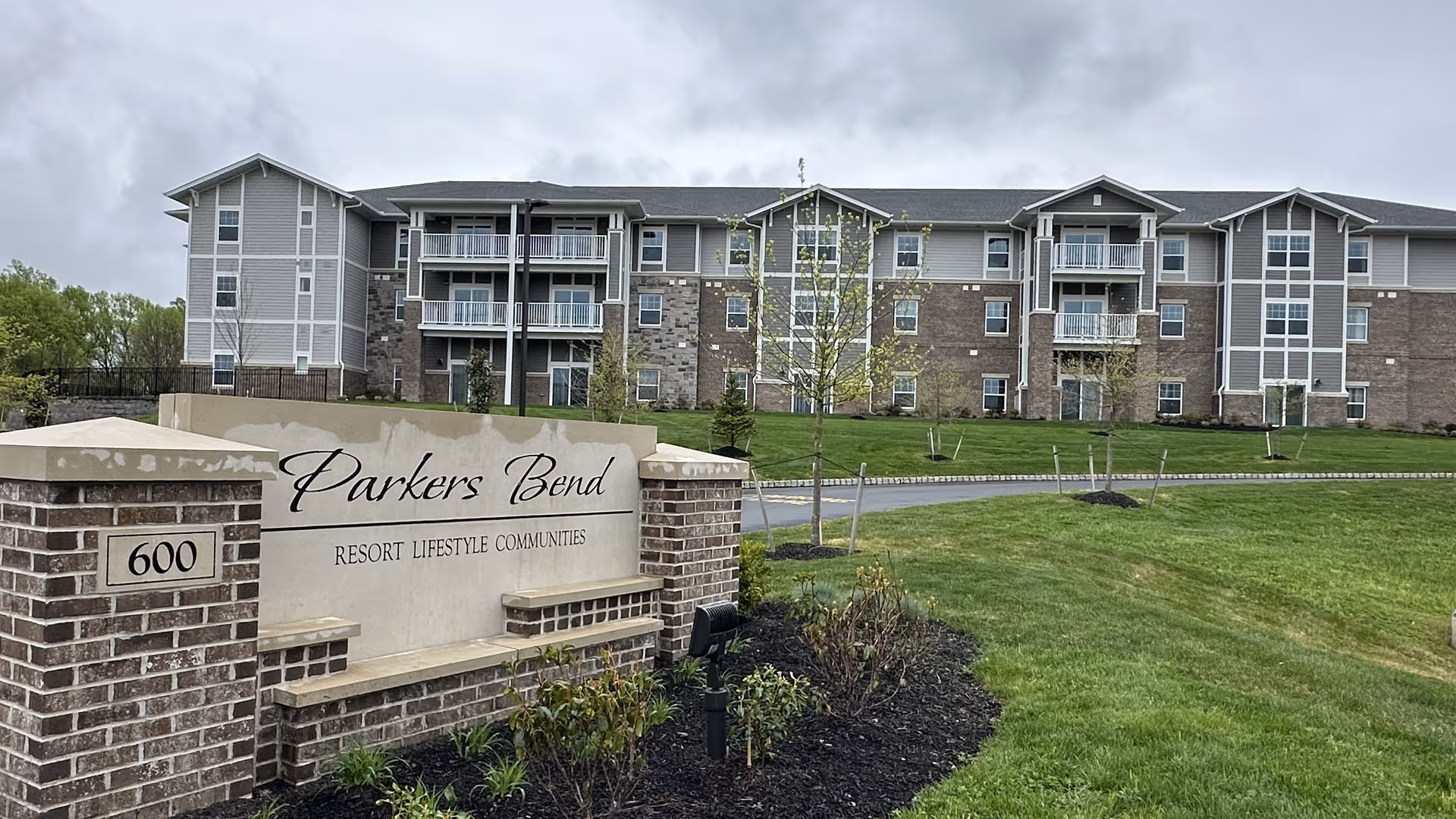 Exterior view of Parkers Bend Retirement Community building with a large sign in the foreground displaying the name and address. The building is multi-story with balconies and a mix of brick and siding exterior. The surrounding area has green grass, small trees, and landscaping under a cloudy sky.
