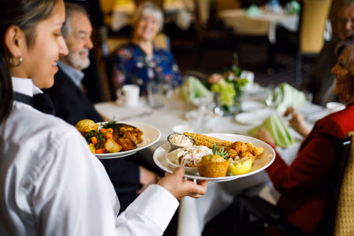 A server carrying two plates of food towards a table where three elderly people are seated, smiling and engaging in conversation in a dining room setting.