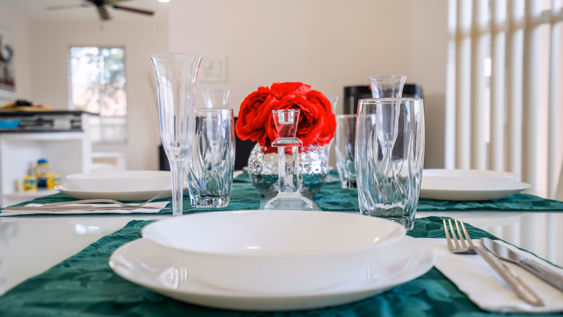 Close-up view of a dining table set with white plates, clear drinking glasses, silverware on white napkins, and a centerpiece of red flowers in a glass vase. The table has a green table runner and the background shows a bright room with windows and a ceiling fan.