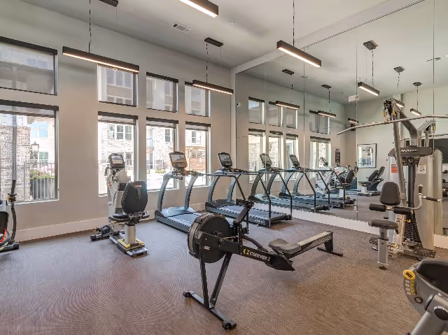 A fitness room with exercise equipment including treadmills, a rowing machine, stationary bikes, and a multi-function weight machine. Large windows let in natural light, and a wall-sized mirror reflects the room.
