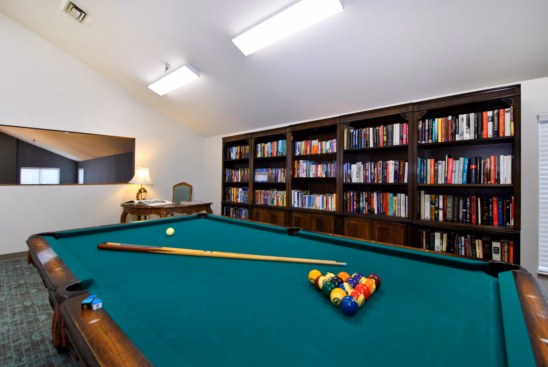 A recreational room featuring a green pool table with a cue stick and a rack of billiard balls set up for a game. Behind the pool table is a large wooden bookshelf filled with books. To the left, there is a small wooden desk with a lamp and a chair. The room has white walls and a carpeted floor.