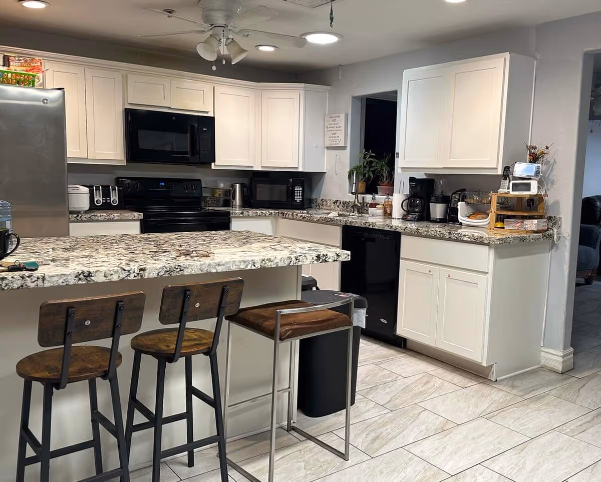 A modern kitchen with white cabinets, granite countertops, and a kitchen island with three wooden bar stools. The kitchen features a stainless steel refrigerator, black microwave, black stove, toaster, coffee maker, and other small appliances. There is a ceiling fan with lights and tiled flooring.