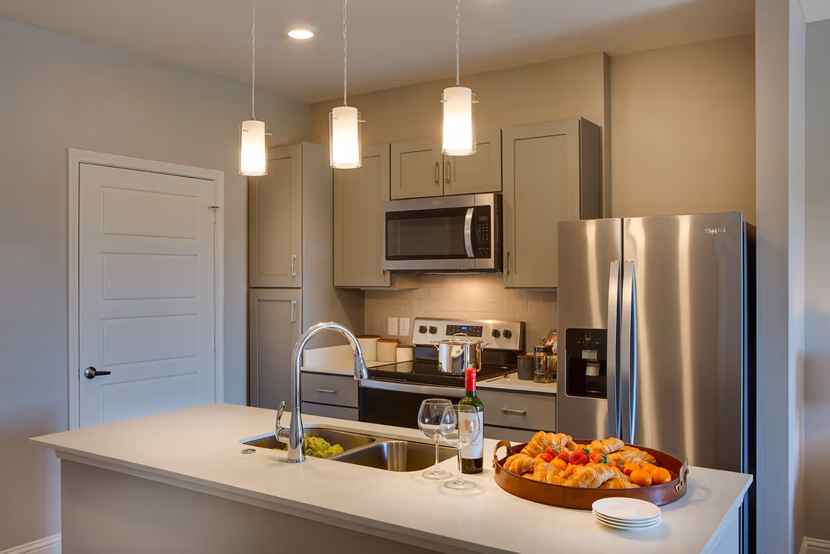 Modern kitchen with gray cabinets, stainless steel refrigerator, microwave, and stove. A white countertop island features a sink, a bottle of wine with two wine glasses, a tray of croissants and fruit, and a stack of small plates. Three pendant lights hang above the island.