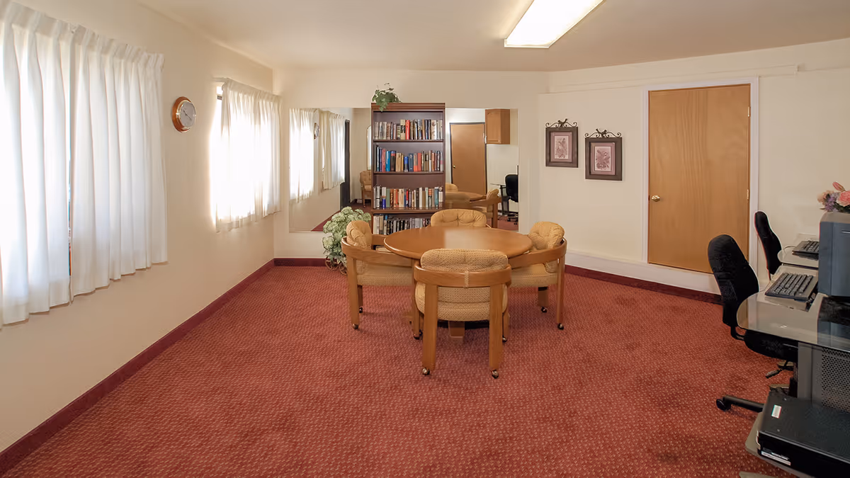 A well-lit room with red carpet flooring featuring a round wooden table surrounded by four cushioned chairs. On the right side, there are two computer workstations with black office chairs. A bookshelf filled with books is positioned against the far wall, next to a large mirror. The room has cream-colored walls, two framed pictures, a wooden door, and windows with white curtains letting in natural light.