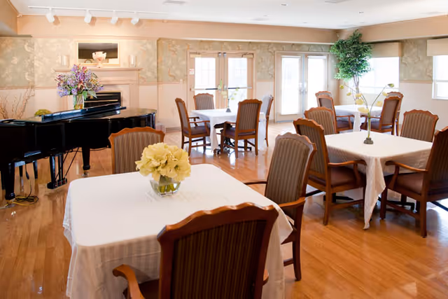 Bright dining room with several tables covered in white cloths, wooden chairs, a grand piano, and floral centerpieces.