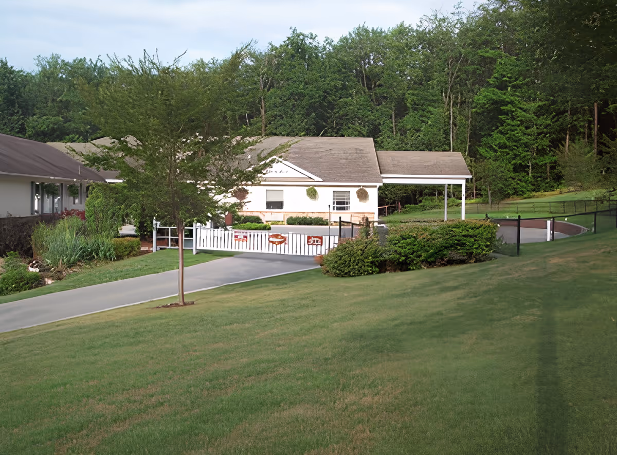 A grassy outdoor area with a paved driveway leading to a white gate and a small white building with a covered porch. The area is surrounded by trees and bushes, creating a peaceful, natural environment.