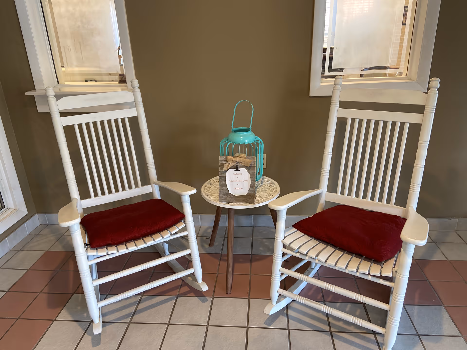 Two white wooden rocking chairs with red cushions are placed on a tiled floor with a small round table between them. On the table, there is a decorative turquoise lantern and a small wooden sign that reads 'Solstice Senior Living at Groton'. The background wall is beige with two windows.