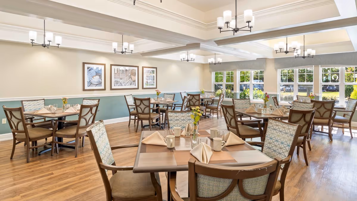 A bright and spacious dining room with multiple wooden tables and chairs arranged neatly. Each table is set with folded napkins, cups, and small flower vases. Large windows on one side allow natural light to fill the room, and framed pictures hang on the opposite wall. The floor is wooden, and modern ceiling lights provide additional illumination.