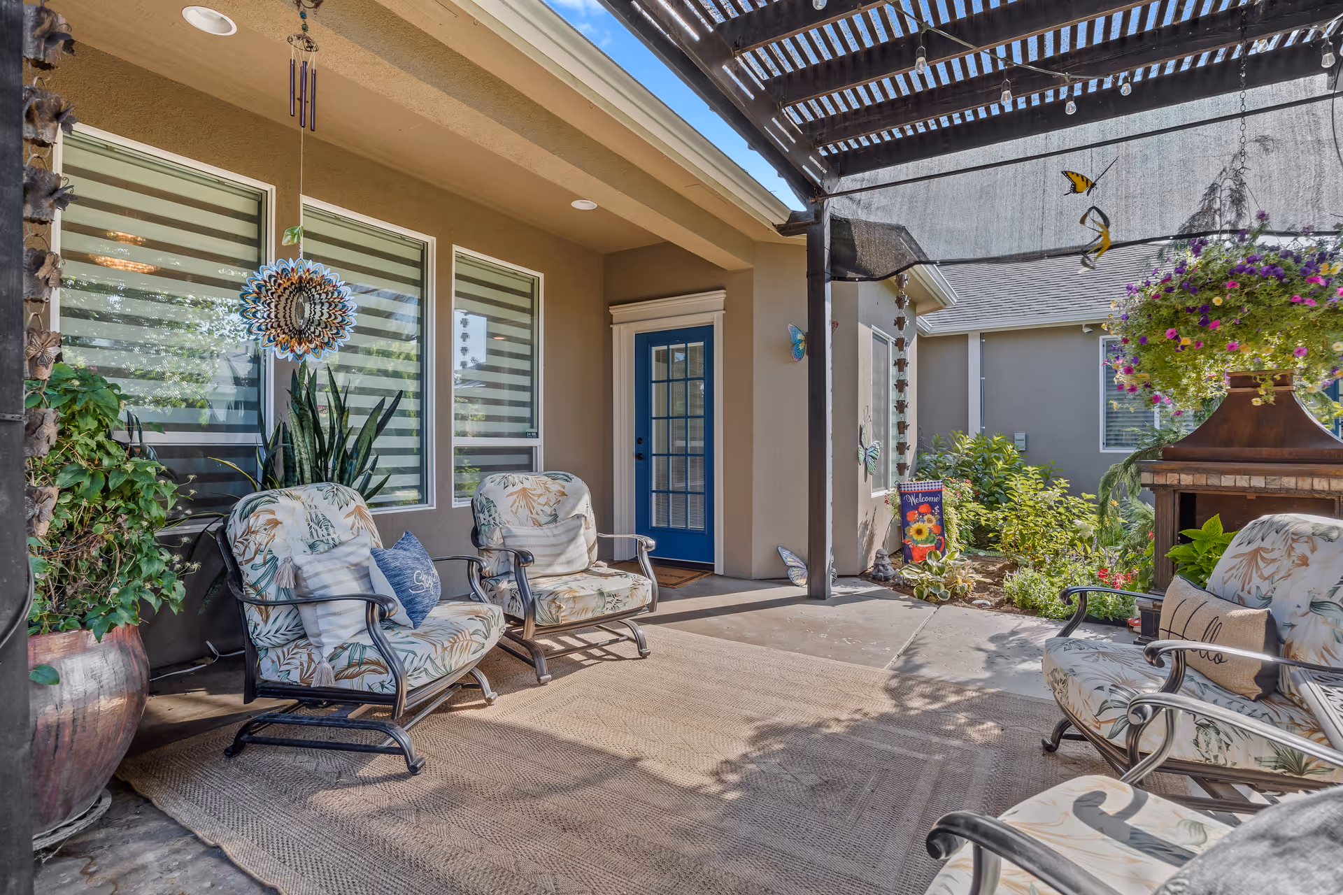 Shaded patio with cushioned metal lounge chairs, potted plants, a hanging flower basket, and a blue door leading into the building.