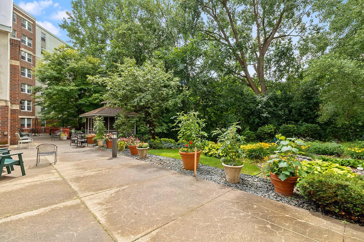Outdoor patio area at Eagle Crest Assisted Living with potted plants lined along the edge, surrounded by lush green trees and shrubs. There are chairs and tables on the concrete patio, and a small gazebo structure is visible in the background next to the building.