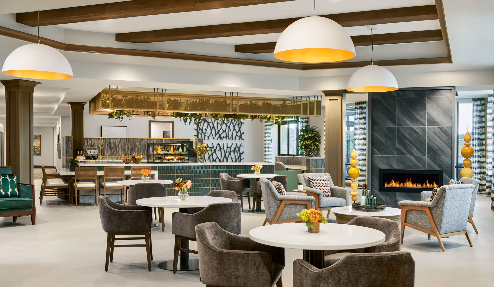 A spacious and modern senior living common area with round tables and cushioned chairs arranged for dining and socializing. The room features a contemporary fireplace with gray tile surround, decorative yellow sculptures on either side, and large pendant lights hanging from a ceiling with wooden beams. In the background, there is a counter area with bar stools and a display case with food items, along with decorative wall art and plants.