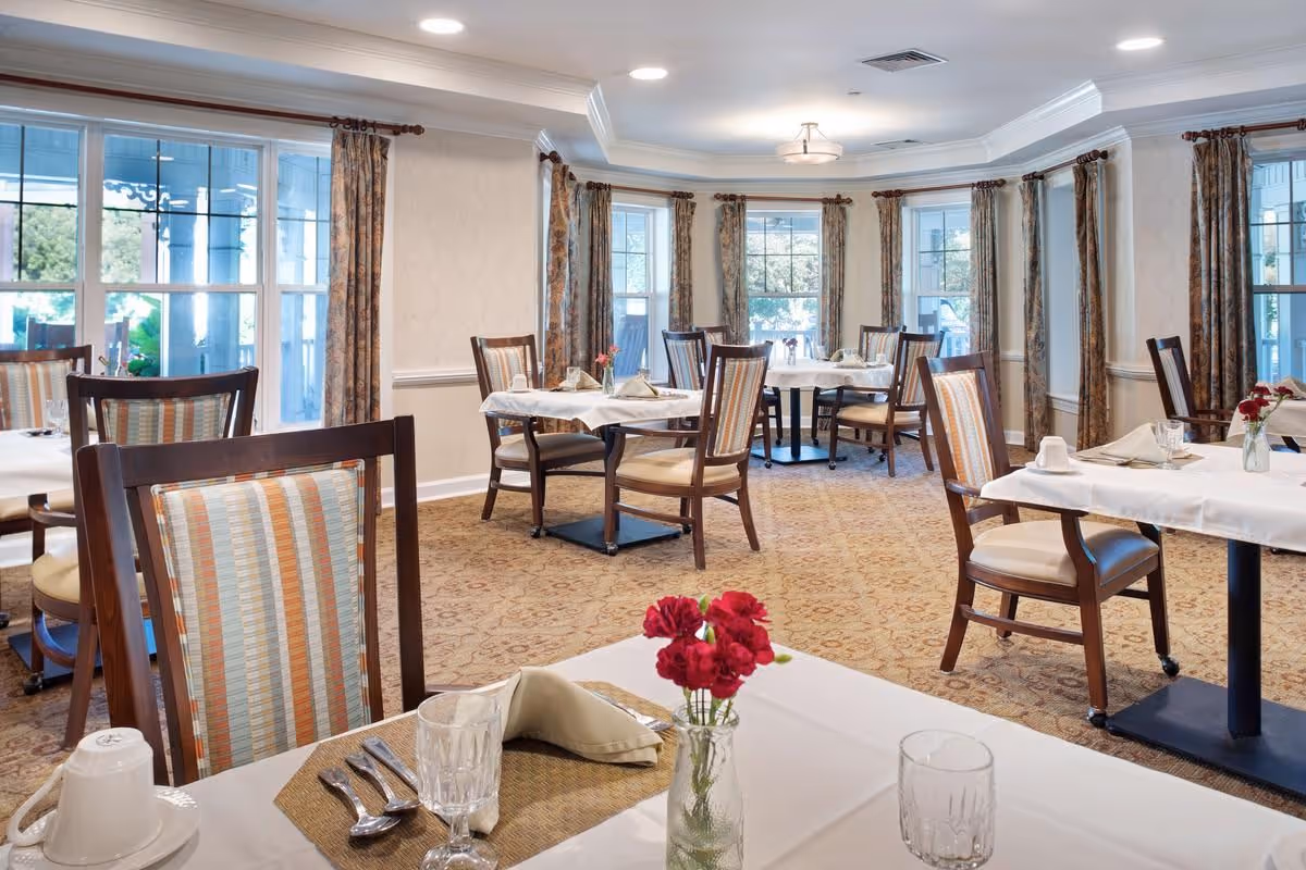 Well-lit dining room with neatly set tables, wooden chairs, and large windows with curtains.