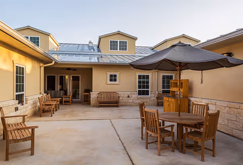 Outdoor courtyard area at Seasons Memory Care at Shavano Park featuring wooden benches and a round wooden table with chairs under a large umbrella, surrounded by beige walls and windows with a metal roof above.