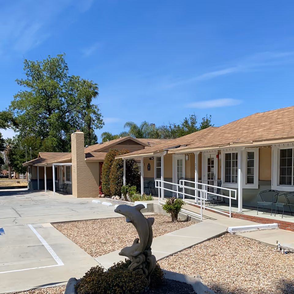 A single-story senior living facility exterior with a front porch, white railing ramp, gravel landscaping, and a dolphin sculpture under a clear blue sky.