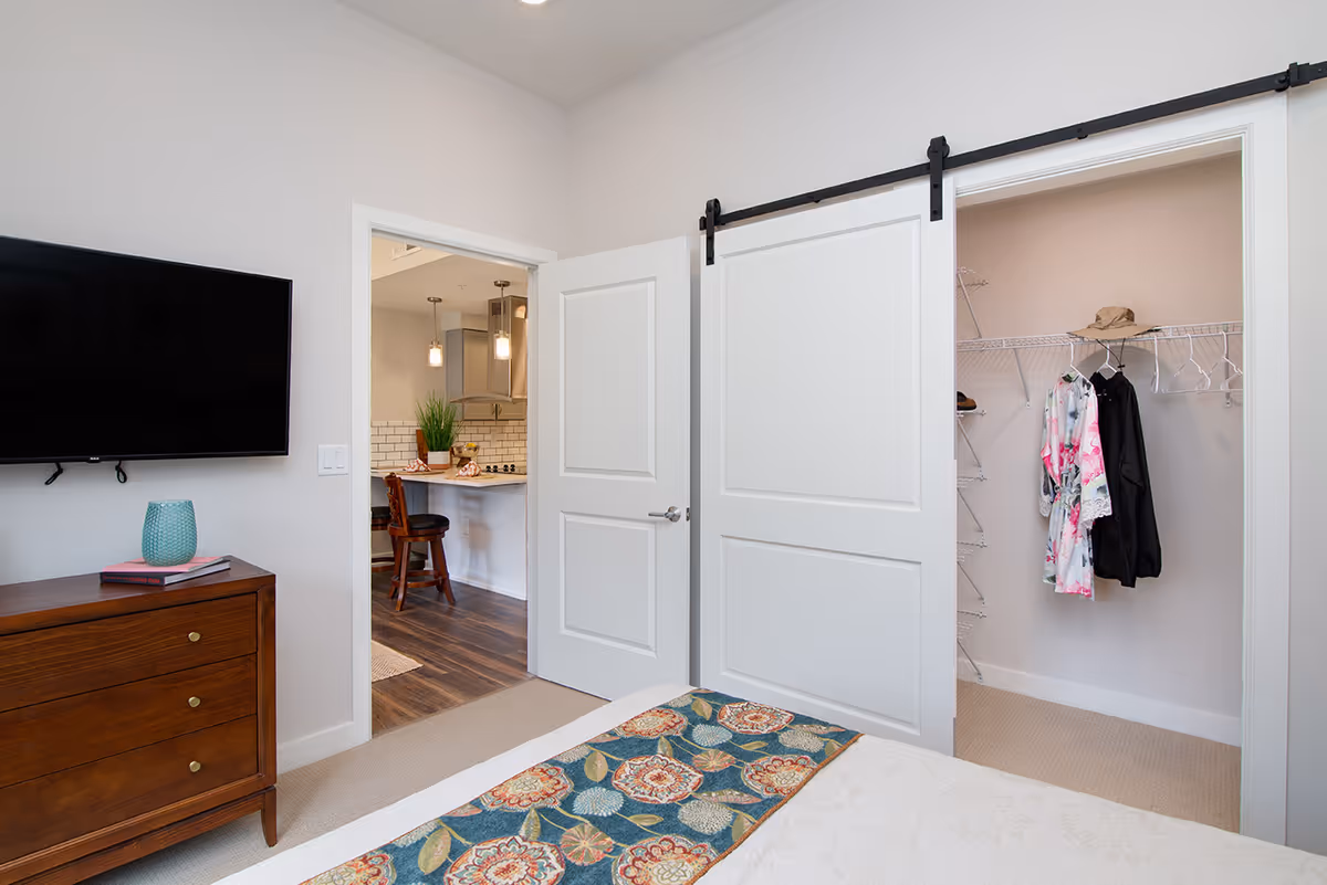 A bedroom with a bed featuring a floral patterned runner, a wooden dresser with a blue vase and books, a wall-mounted flat screen TV, and an open door leading to a kitchen area. There is also a sliding barn door partially open revealing a closet with hanging clothes and shelves.
