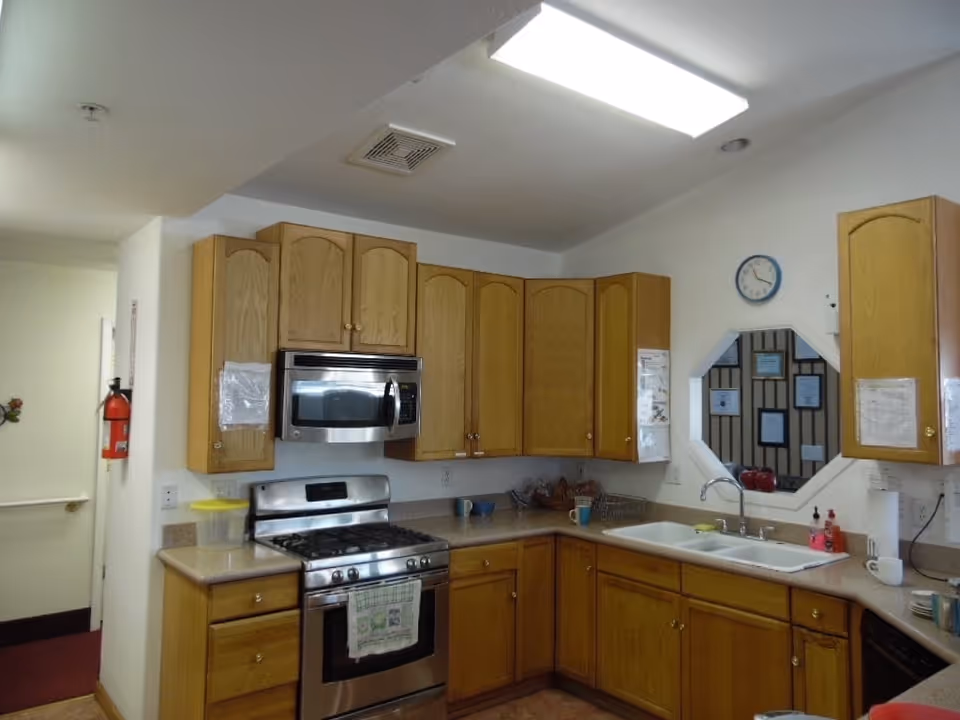 A kitchen area with wooden cabinets, a stainless steel stove and microwave, a double sink, and various kitchen items on the countertops. There is a clock on the wall above the sink and a fire extinguisher mounted on the wall near the entrance.