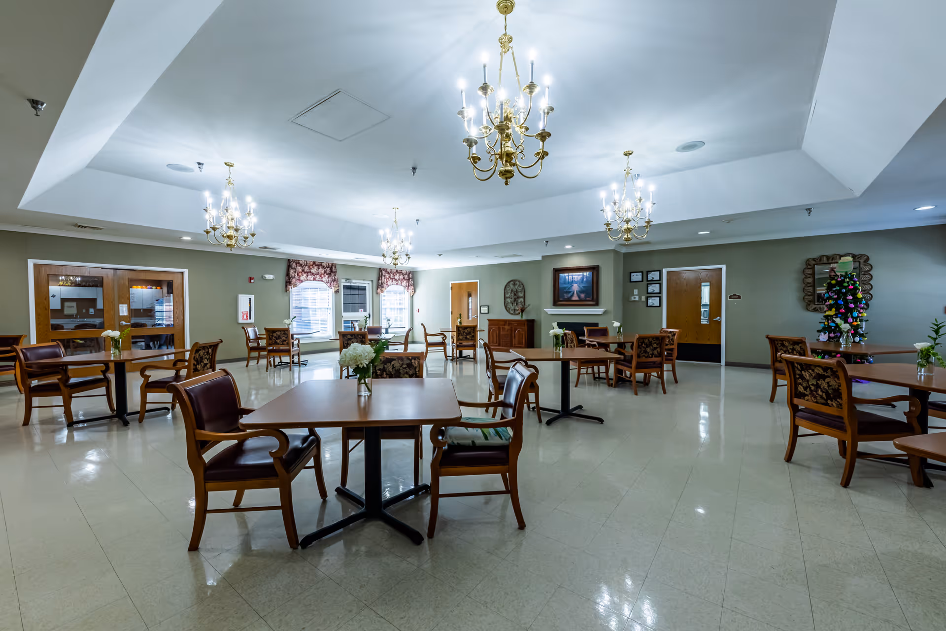 A spacious dining room with multiple wooden tables and chairs arranged neatly. The room features a high ceiling with elegant chandeliers providing bright lighting. There are windows with floral curtains, a decorative mirror, framed artwork, and a small decorated Christmas tree in the corner. The floor is tiled and the walls are painted in a muted green color.