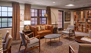 Cozy common room with a leather sofa, armchairs, coffee tables, shelving and large windows.