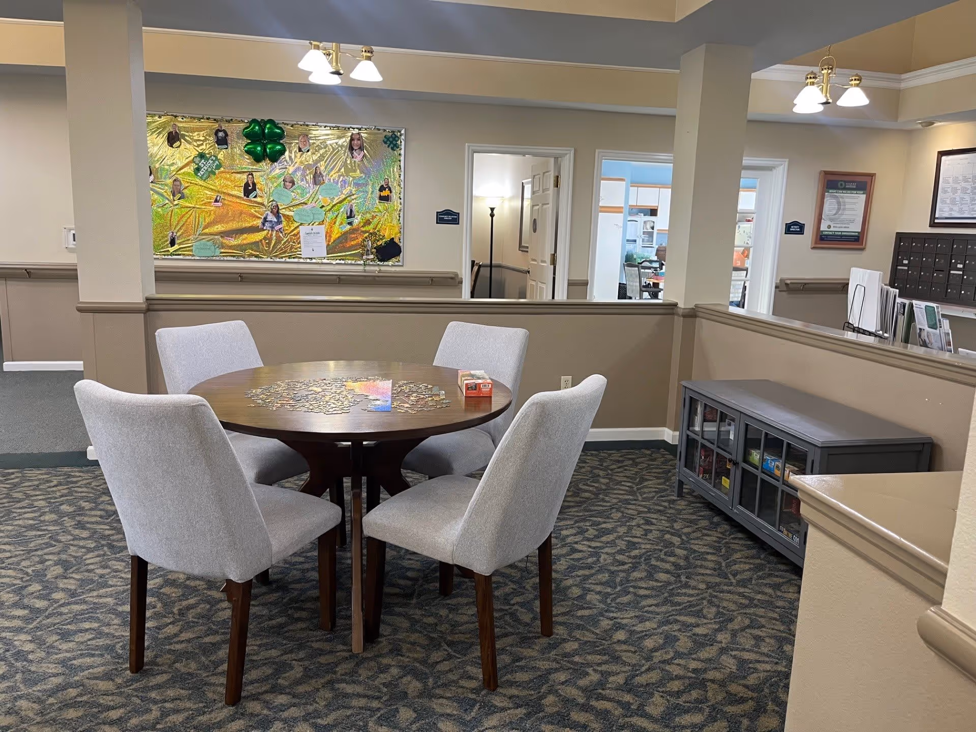 A cozy common area with a round wooden table and four light gray upholstered chairs. A partially completed jigsaw puzzle and a box of puzzle pieces are on the table. The room has patterned carpet, beige walls with a chair rail, and a bulletin board decorated with photos and green decorations. There are doorways leading to other rooms and a cabinet with glass doors containing games or books.
