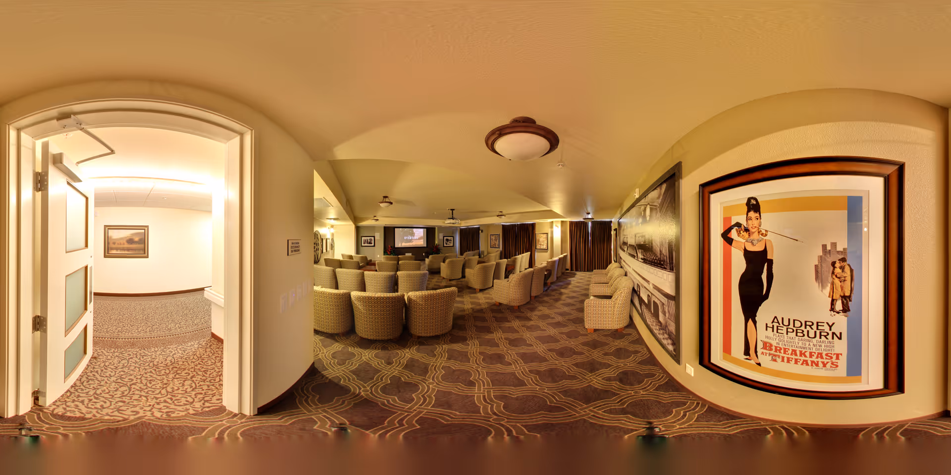 A cozy theater-style room with multiple rows of cushioned chairs facing a large screen. The room has warm lighting and carpeted floors with a patterned design. On the right wall, there is a framed poster of the movie 'Breakfast at Tiffany's' featuring Audrey Hepburn. To the left, an open door leads to a hallway with similar carpet and a framed picture on the wall.