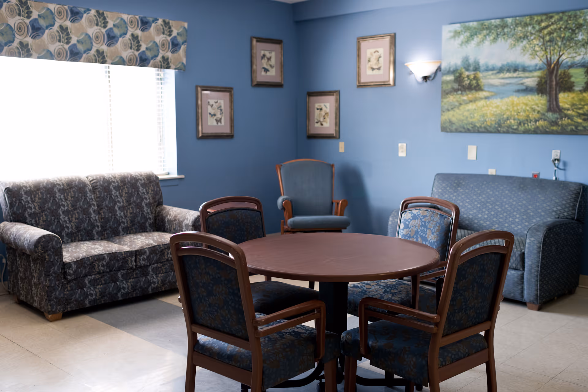 A cozy common area with blue walls featuring a round wooden table surrounded by four upholstered chairs. There are two patterned sofas and a single armchair against the walls. The room is decorated with framed pictures and a large landscape painting. A window with a floral valance allows natural light to enter.