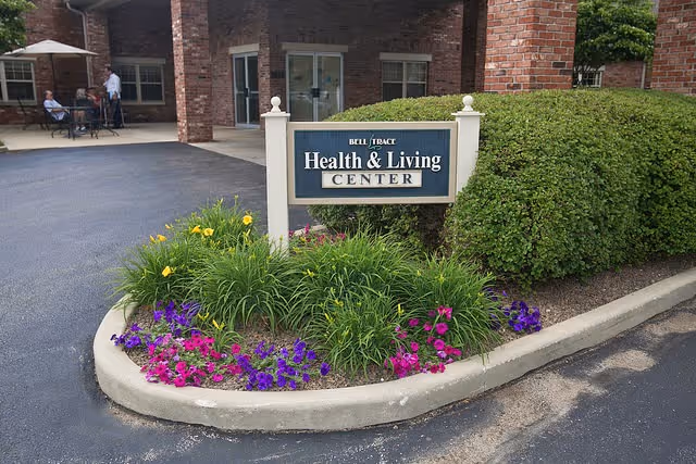 Entrance area of Bell Trace Health & Living Center with a sign displaying the facility name surrounded by a flower bed with purple and pink flowers and green shrubs. In the background, there is a brick building with windows and a covered patio where a few people are seated at a table.