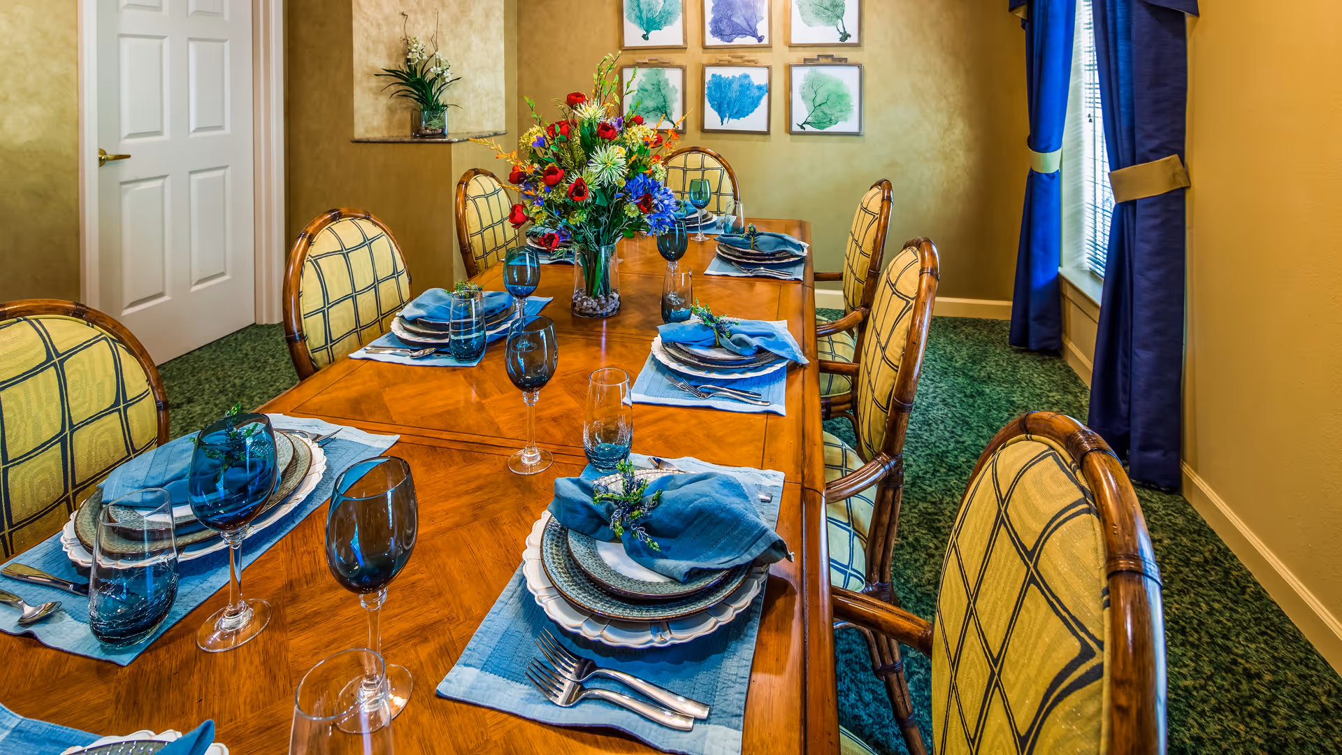 A formal dining room with a long wooden table set for six people. Each place setting includes blue placemats, blue napkins, white plates, and blue glassware. A colorful floral centerpiece is in the middle of the table. The room has green carpet, yellow walls, a window with blue curtains, and framed artwork on the wall.