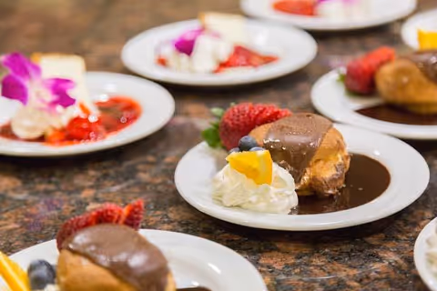 Close-up view of dessert plates on a granite countertop, each plate featuring a chocolate-covered pastry, whipped cream topped with a small piece of orange, and fresh strawberries and blueberries, with some plates also garnished with a purple edible flower.