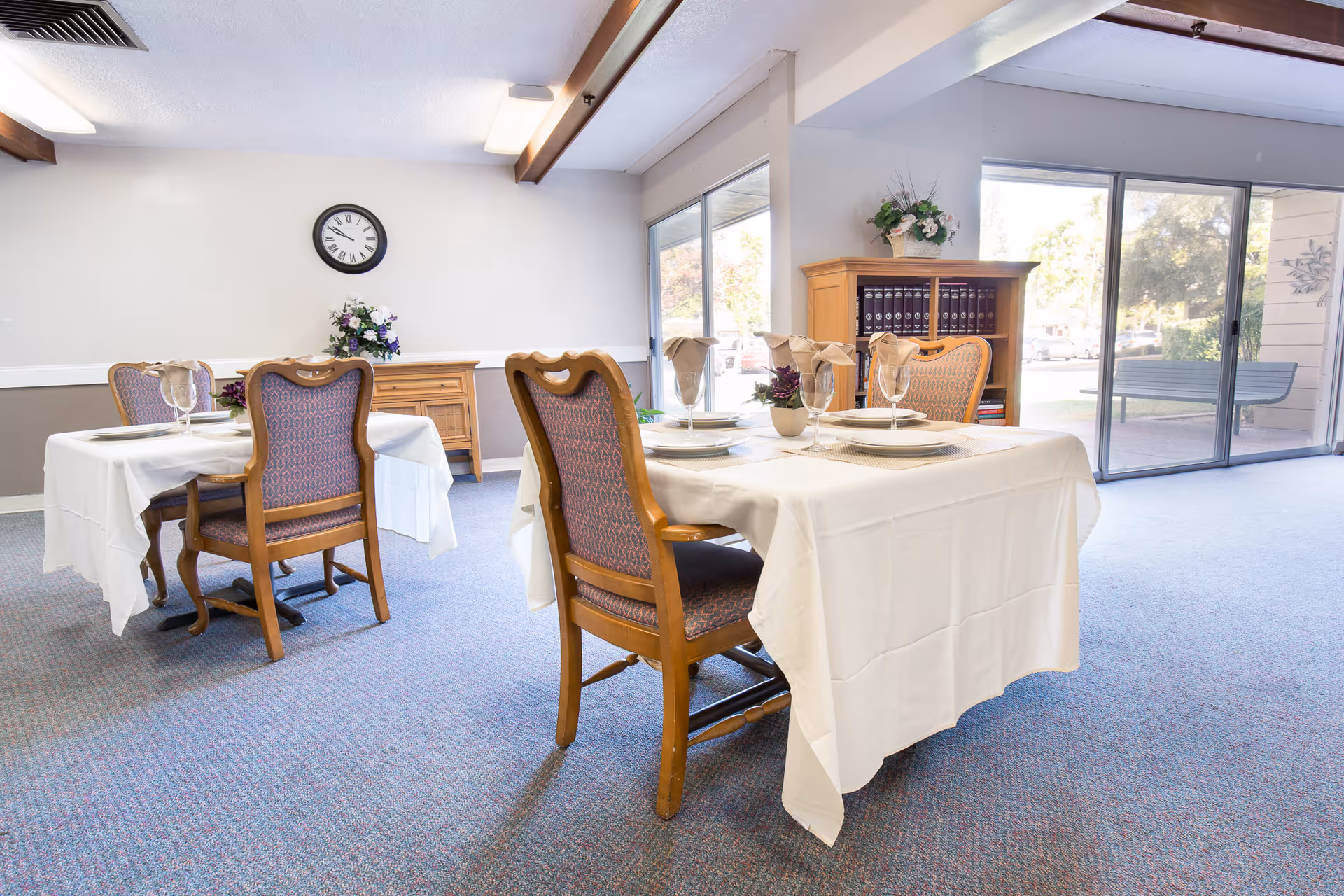 A bright dining room with two tables covered in white tablecloths, each set with plates, glasses, and folded napkins. The room has carpeted flooring, a clock on the wall, a wooden cabinet with books and flowers, and large sliding glass doors leading outside.