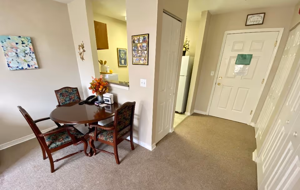 A small dining area with a round wooden table and four upholstered chairs with floral patterns. The table has a flower arrangement and some small items on it. To the right, there is a hallway with a white door and closets with white louvered doors. A kitchen area with a refrigerator is visible through a pass-through window and an open doorway. The walls are light-colored, and there is a floral painting on the left wall.