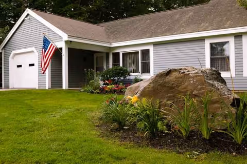Single-story senior living facility exterior with American flag, manicured lawn, and landscaped rock garden.