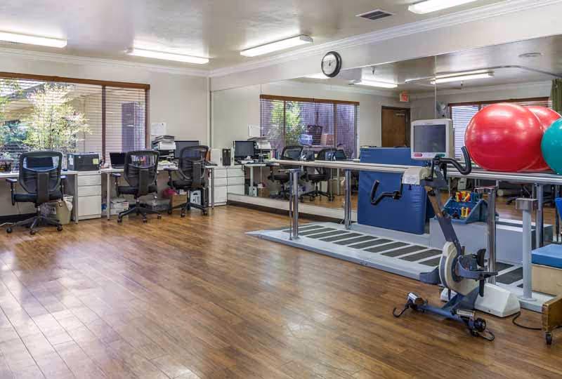 A physical therapy or rehabilitation room with wooden flooring, several office chairs and desks with computers along the left wall, large mirrors on the back wall, and exercise equipment including a stationary bike and large exercise balls on the right side.