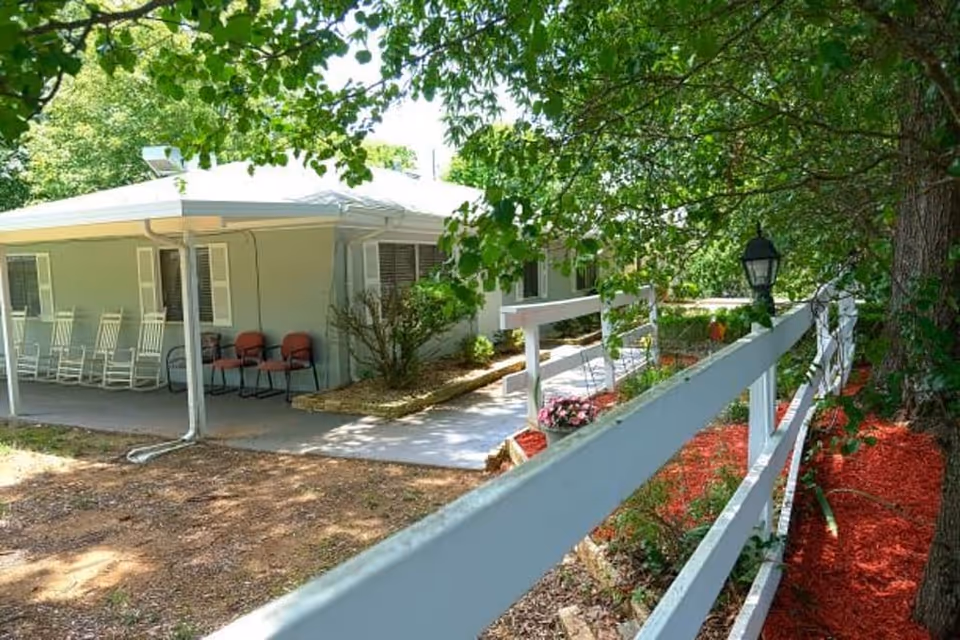 Outdoor view of a senior living facility with a white fence, a pathway, and a covered porch area with several chairs. The area is surrounded by trees and landscaping with red mulch.