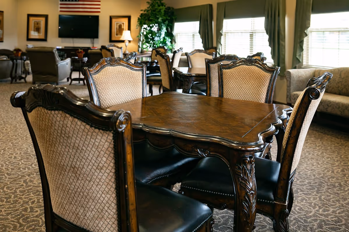 A cozy senior living common area with ornate wooden tables and upholstered chairs arranged on a patterned carpet. The room has large windows with curtains allowing natural light, a TV mounted on the wall beneath an American flag, framed pictures, a lamp, and a green potted plant in the background.