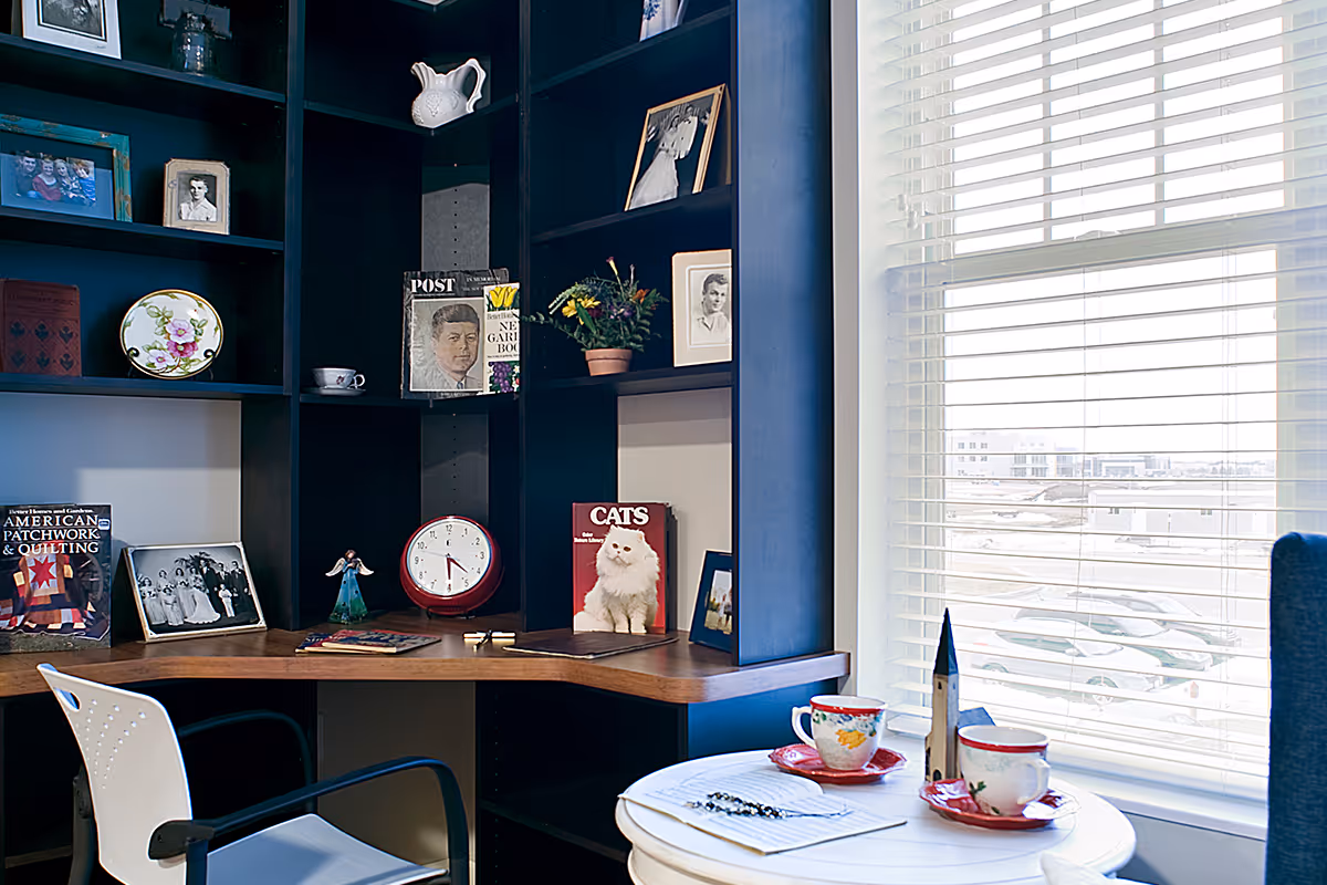 A cozy corner workspace with dark blue built-in shelves filled with framed photos, books, a clock, and decorative items. A white chair is positioned at a wooden desk. Next to the desk is a small round table with two floral teacups and saucers, a small model of a church, and a notebook with a pen. A large window with white blinds lets in natural light and shows a snowy outdoor parking area.